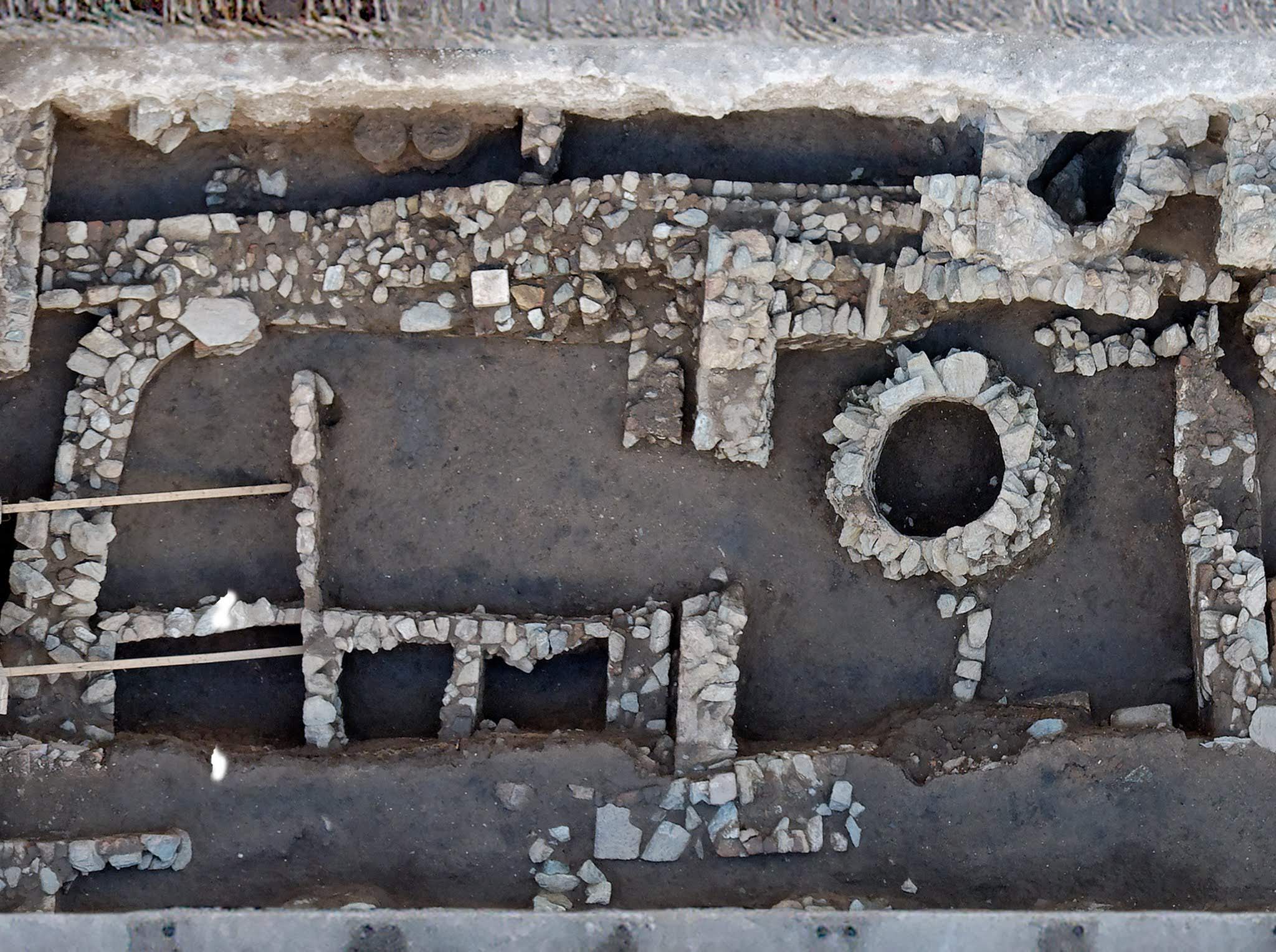 A high-angle overhead view of an archaeological excavation site, detailing ancient stone foundations, partitioned chambers, and a circular stone well structure.