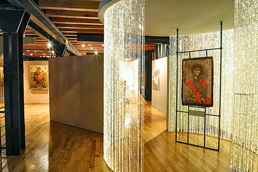 A modern museum interior showcasing ancient Byzantine religious icons, accented by shimmering, illuminated string curtains and warm gallery lighting.