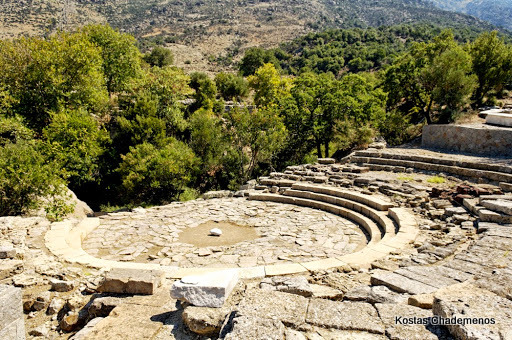 An ancient stone semi-circular theater or council chamber (bouleuterion) situated amidst lush green vegetation and hillsides under bright daylight.