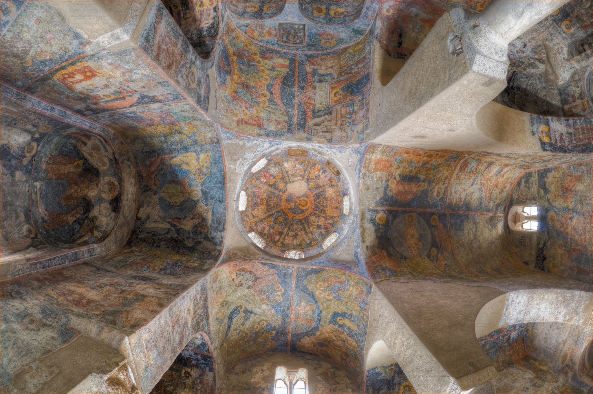 A low-angle view looking directly up at the vaulted ceiling and central dome of an ancient Christian church, covered in detailed Byzantine frescoes.
