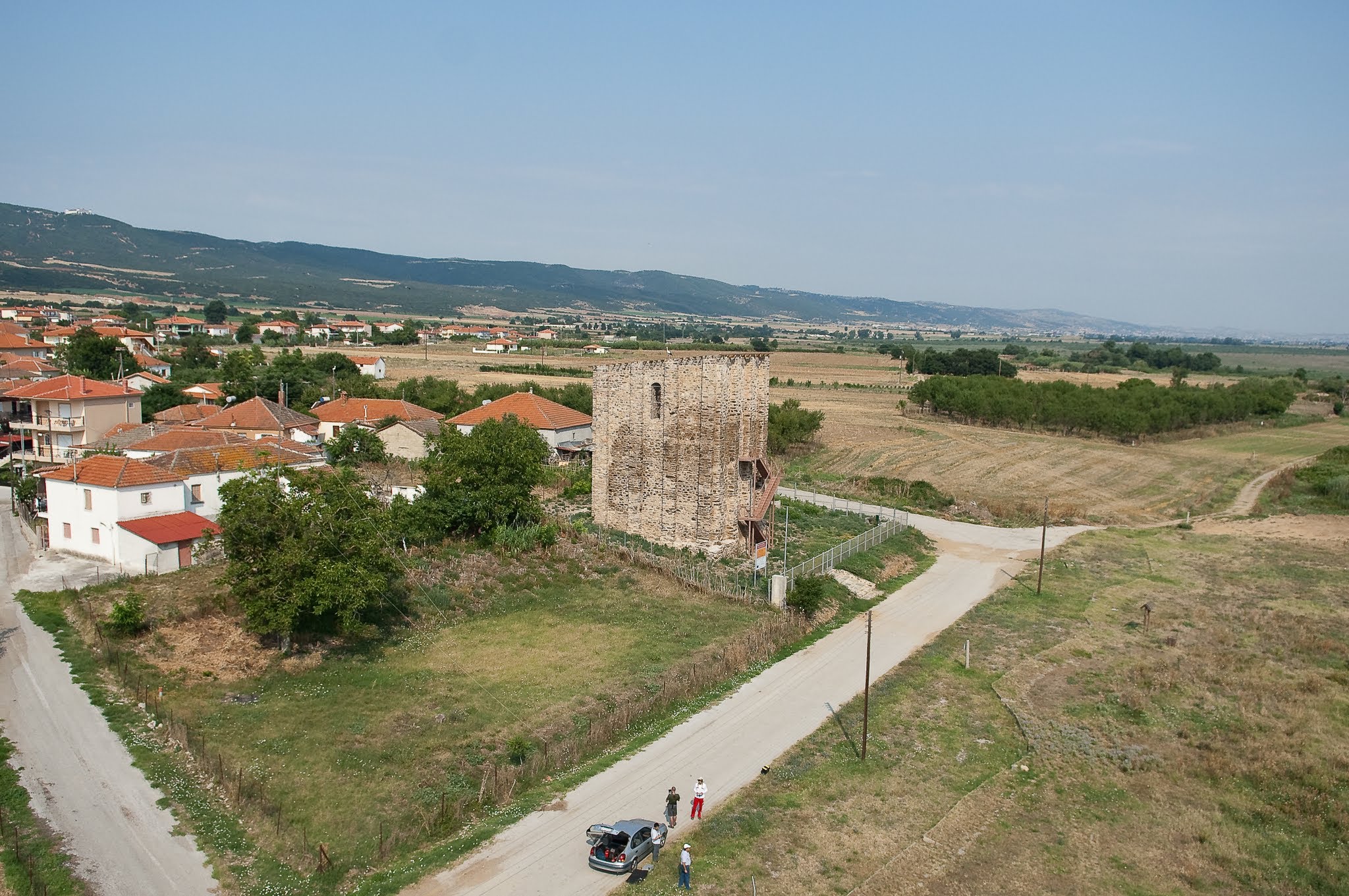 An aerial view of the Byzantine Tower of Marianos, showing the surrounding countryside, a village edge, and the flight crew parked along a dirt road.
