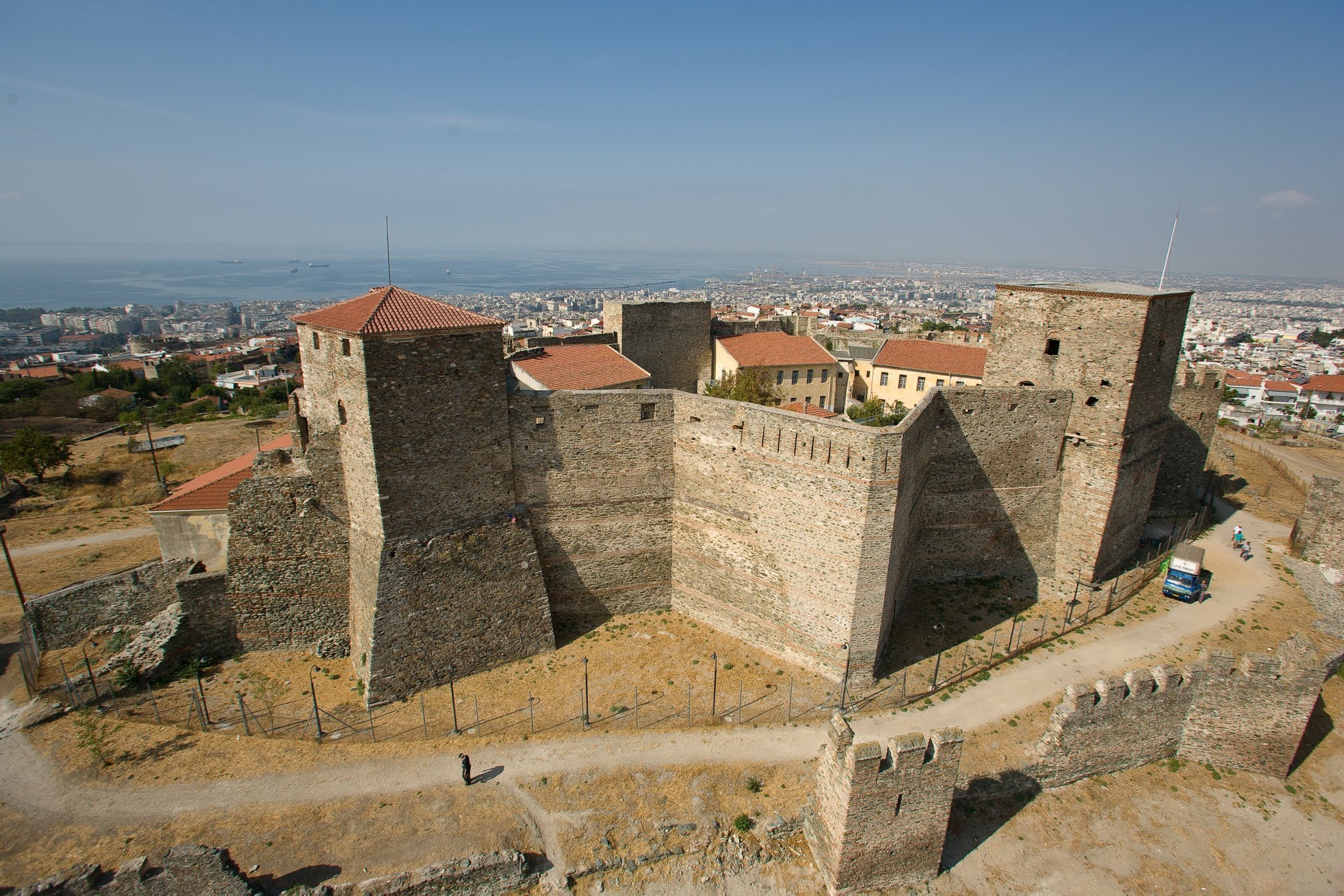 An elevated view of the Heptapyrgion fortress in Thessaloniki, showcasing its defensive walls, tiled-roof towers, and the sprawling city and Thermaic Gulf in the background.