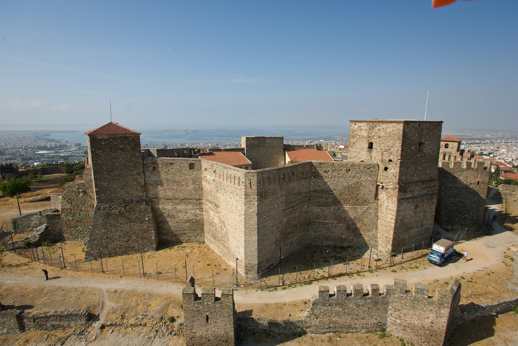 An elevated wide-angle view of the Heptapyrgion fortress in Thessaloniki, showcasing its massive stone walls, central towers, and the sprawling city descending toward the sea.