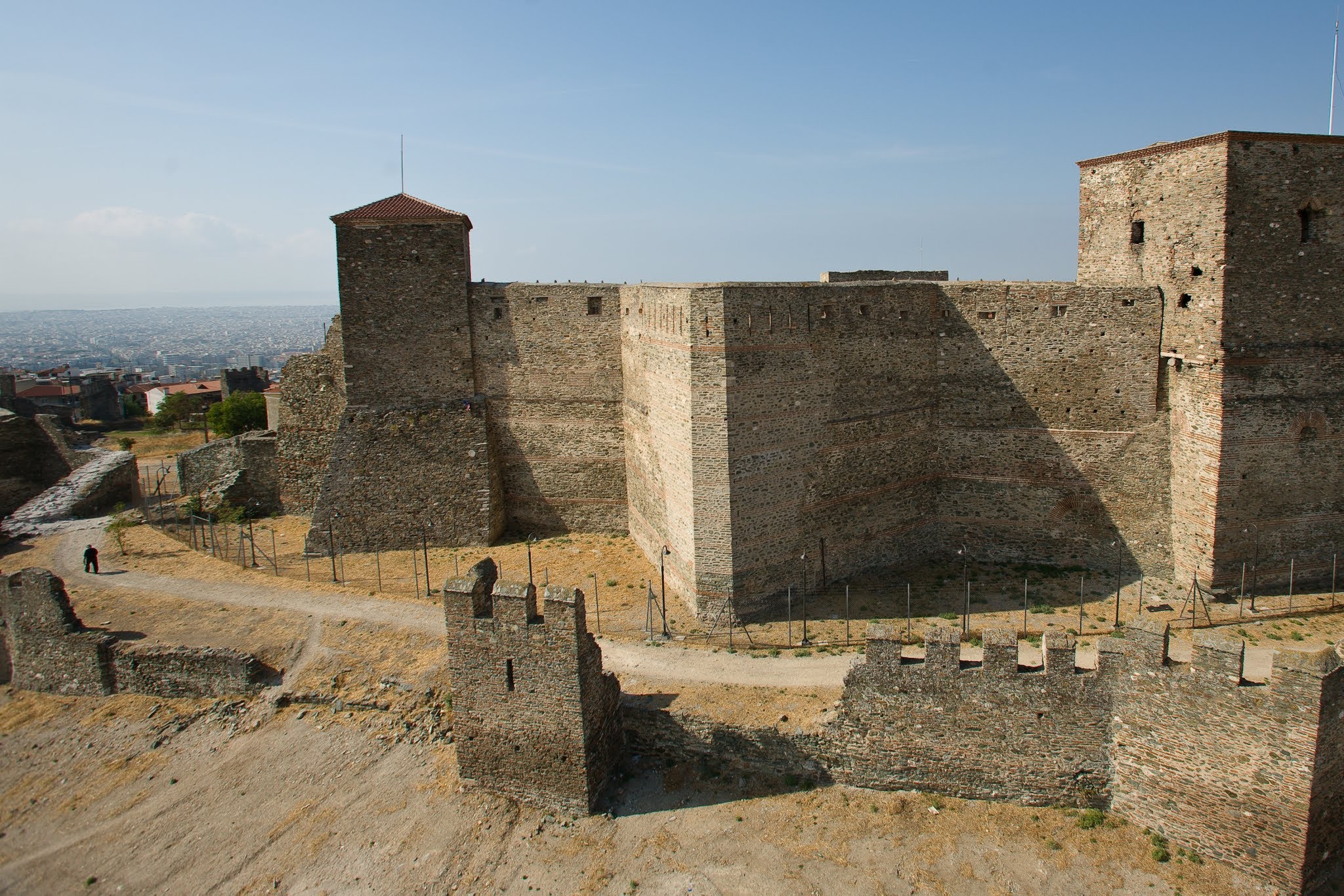 An elevated medium-wide shot of the Heptapyrgion fortress walls and towers, showcasing the massive stone masonry and defensive structures under clear daylight.