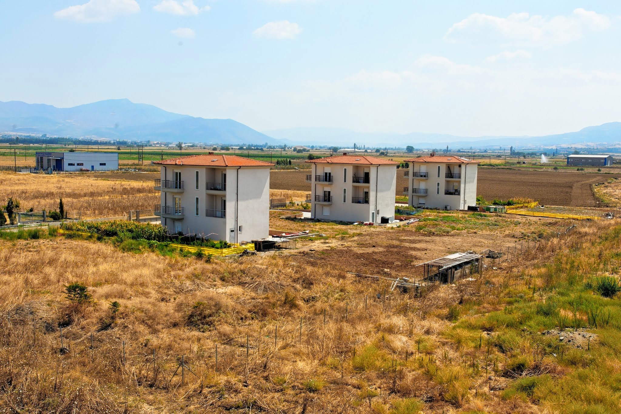An aerial view of three identical, newly built modern three-story apartment buildings standing in a dry, grassy field against a backdrop of distant mountains.