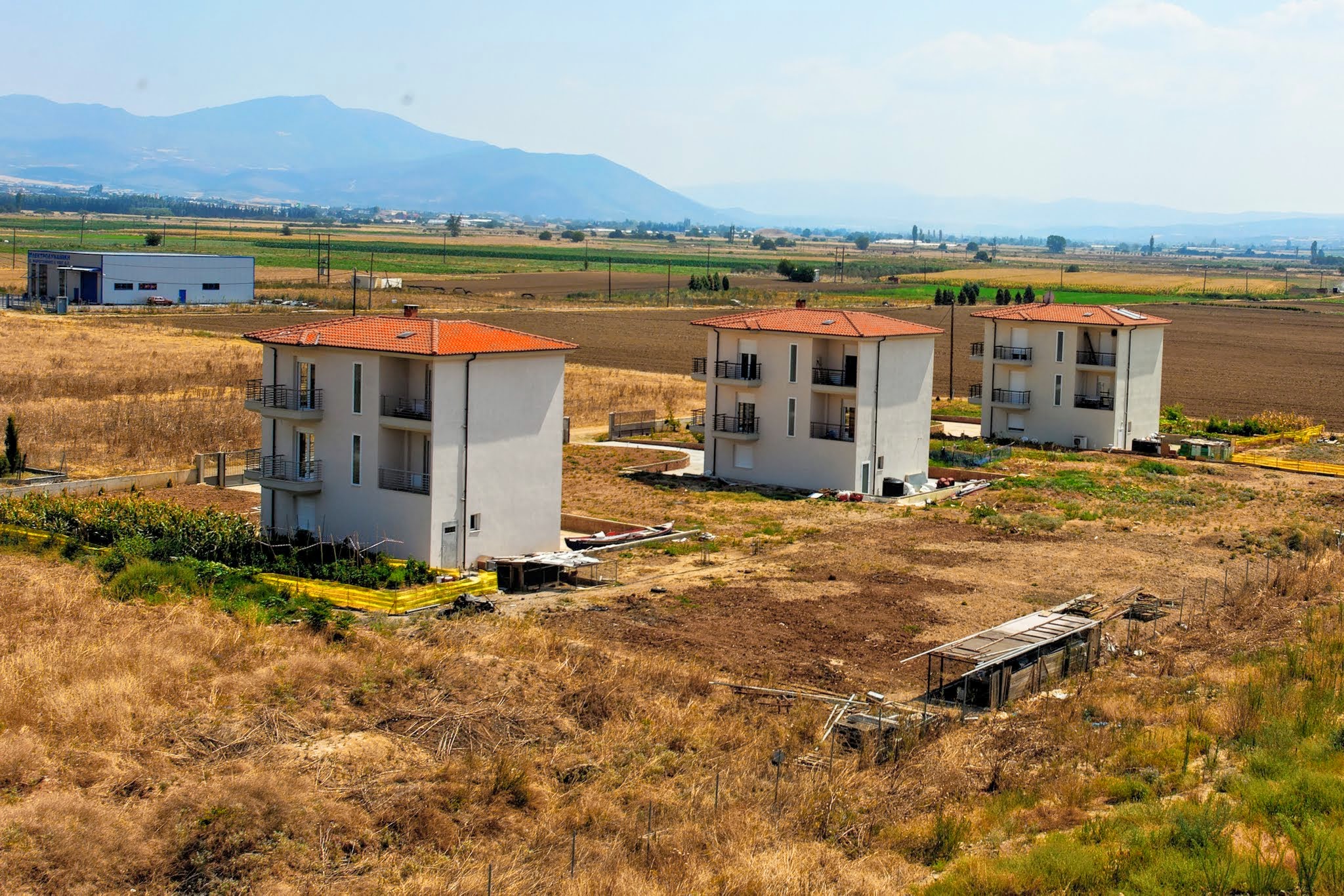 A close-up aerial view of three modern three-story houses standing in a line in a dry rural landscape, with agricultural fields and distant hills in the background.
