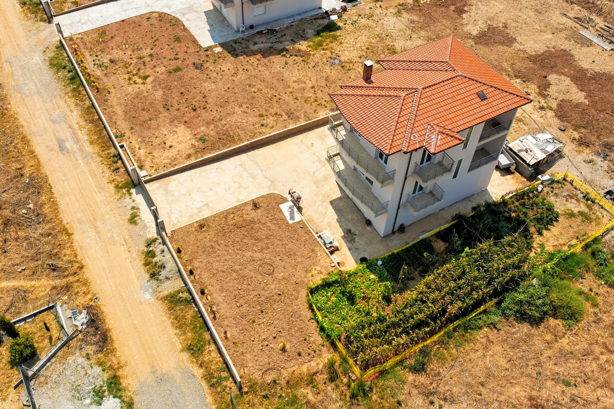 A top-down, high-angle aerial view of a three-story residential house, detailing its red tile roof, concrete courtyard, enclosed yard, and adjacent garden.