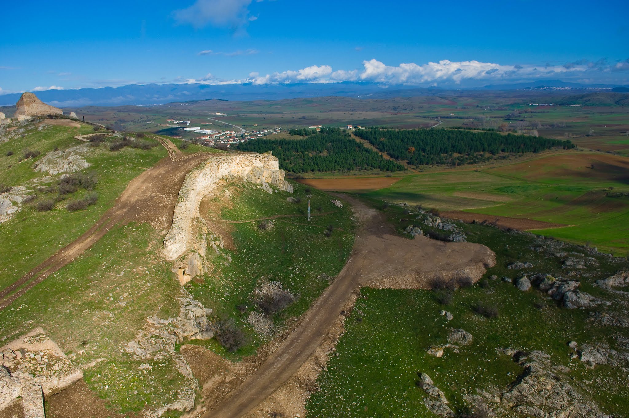 High-angle aerial view of historic stone ruins on a grassy hillside, overlooking a rolling countryside with forests and distant mountains.