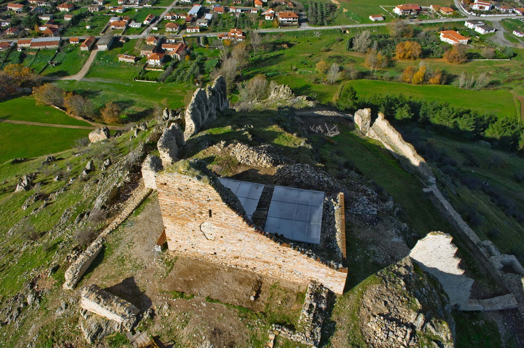 Aerial view looking down at ancient brick and stone fortress ruins on a hilltop, with a modern protective roof and a village in the background.