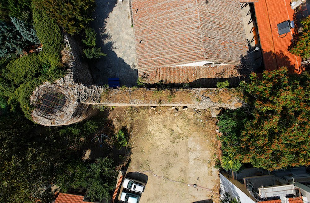 A top-down aerial view of a section of Byzantine stone wall, featuring an ivy-draped circular tower, adjacent church roof, and urban courtyard in Thessaloniki.