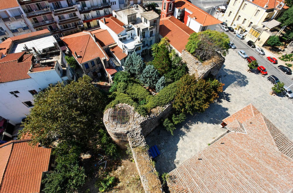 An elevated high-angle view of a section of Thessaloniki's old city walls, showing a round tower covered in ivy, adjacent buildings, and streets with parked cars.