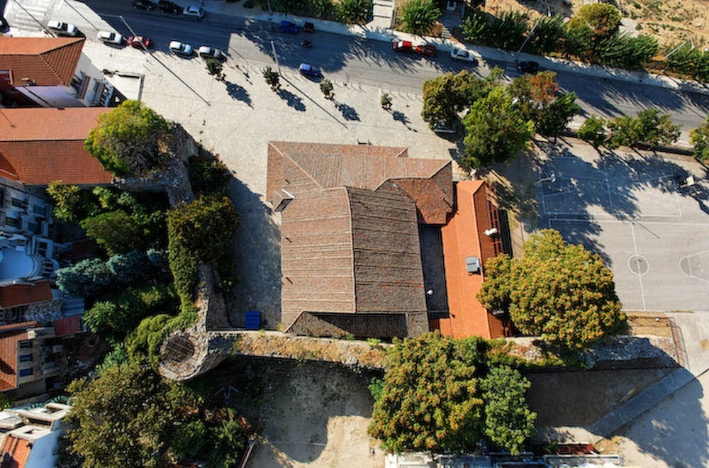 A top-down aerial view of a Byzantine wall segment and a church with a tiled roof, next to a paved sports court and a street in Thessaloniki.
