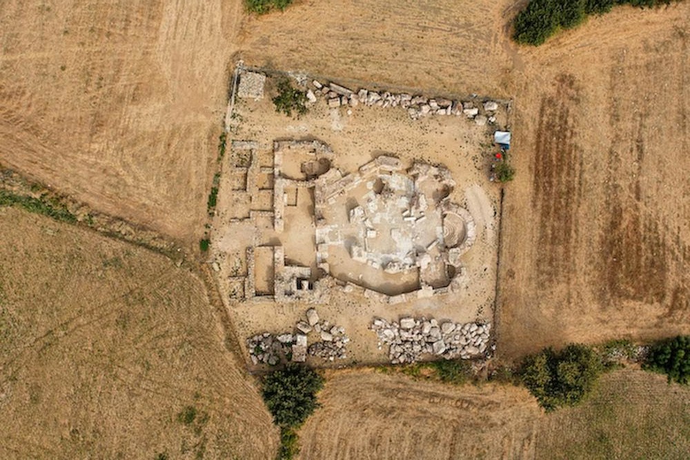A top-down aerial photograph of the archaeological site of an ancient basilica, detailing its geometric layout, foundations, and surrounding fields.