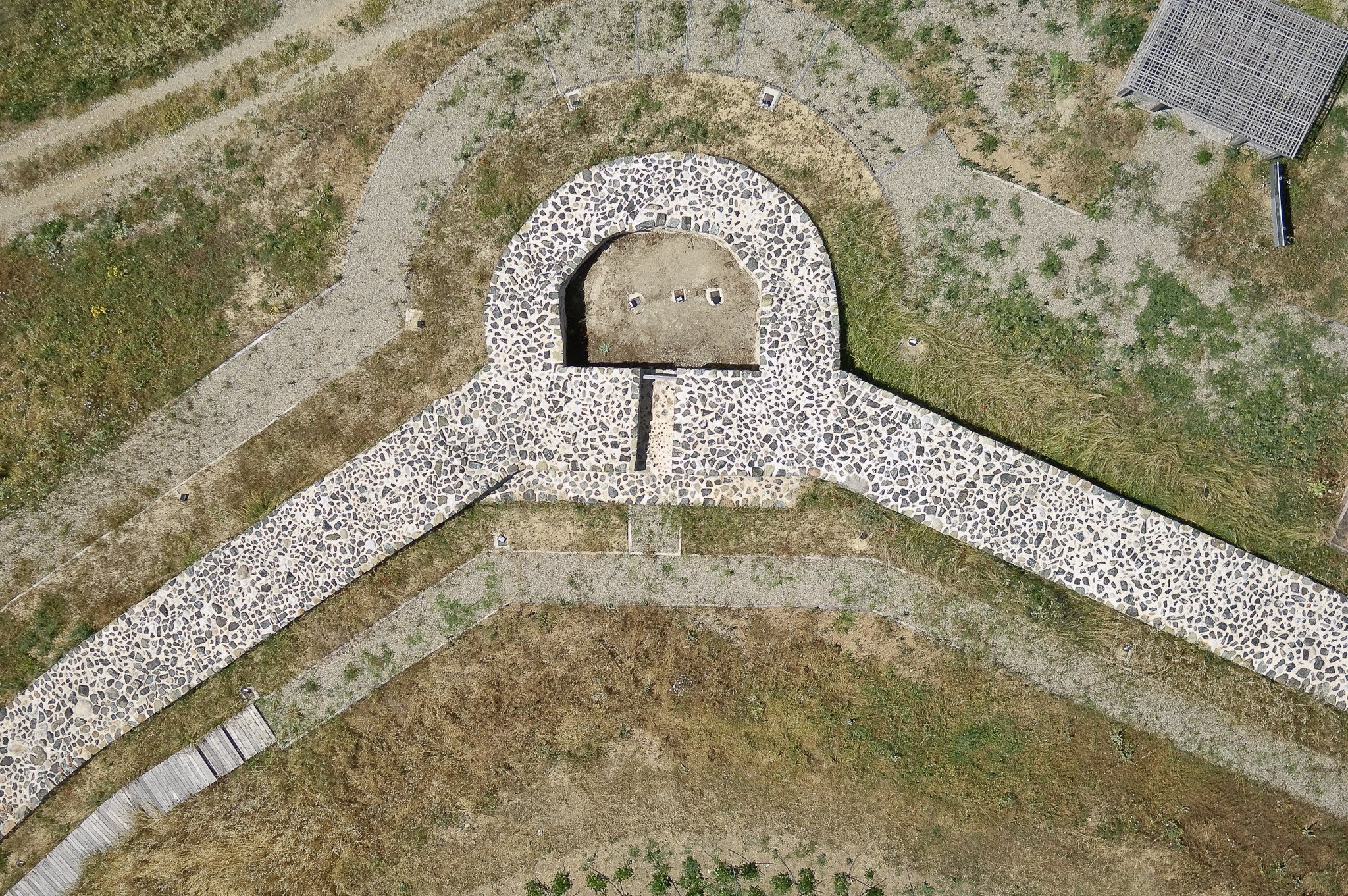Symmetrical top-down aerial photograph of a historic site showing stone foundations, circular ruins, gravel paths, and visitor platforms.