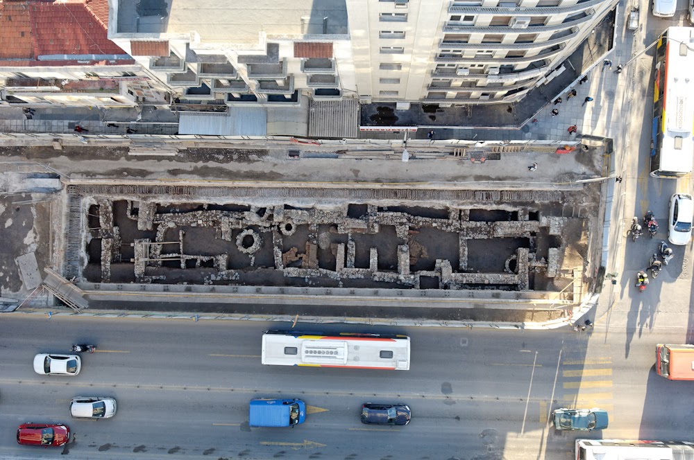 A top-down aerial photograph of an active archaeological excavation site situated between busy urban roadways and high-rise buildings. The ruins reveal ancient stone foundation walls and structural footprints exposed during city development.