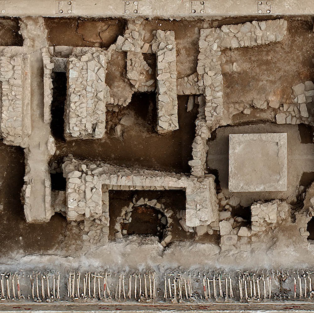 A top-down detailed aerial photograph of excavated archaeological ruins, showcasing ancient stone wall foundations, a square plinth, and a semi-circular stone feature.