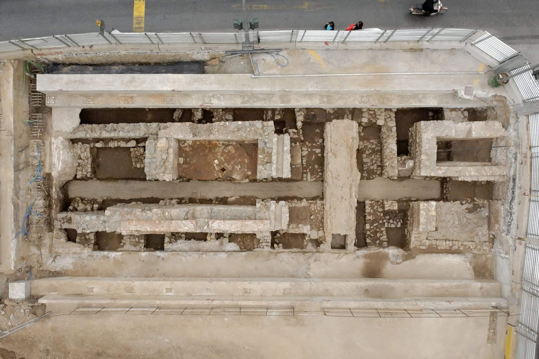 Nadir aerial photograph of an urban archaeological excavation site, showing stone ruins surrounded by modern concrete retaining walls and fencing.