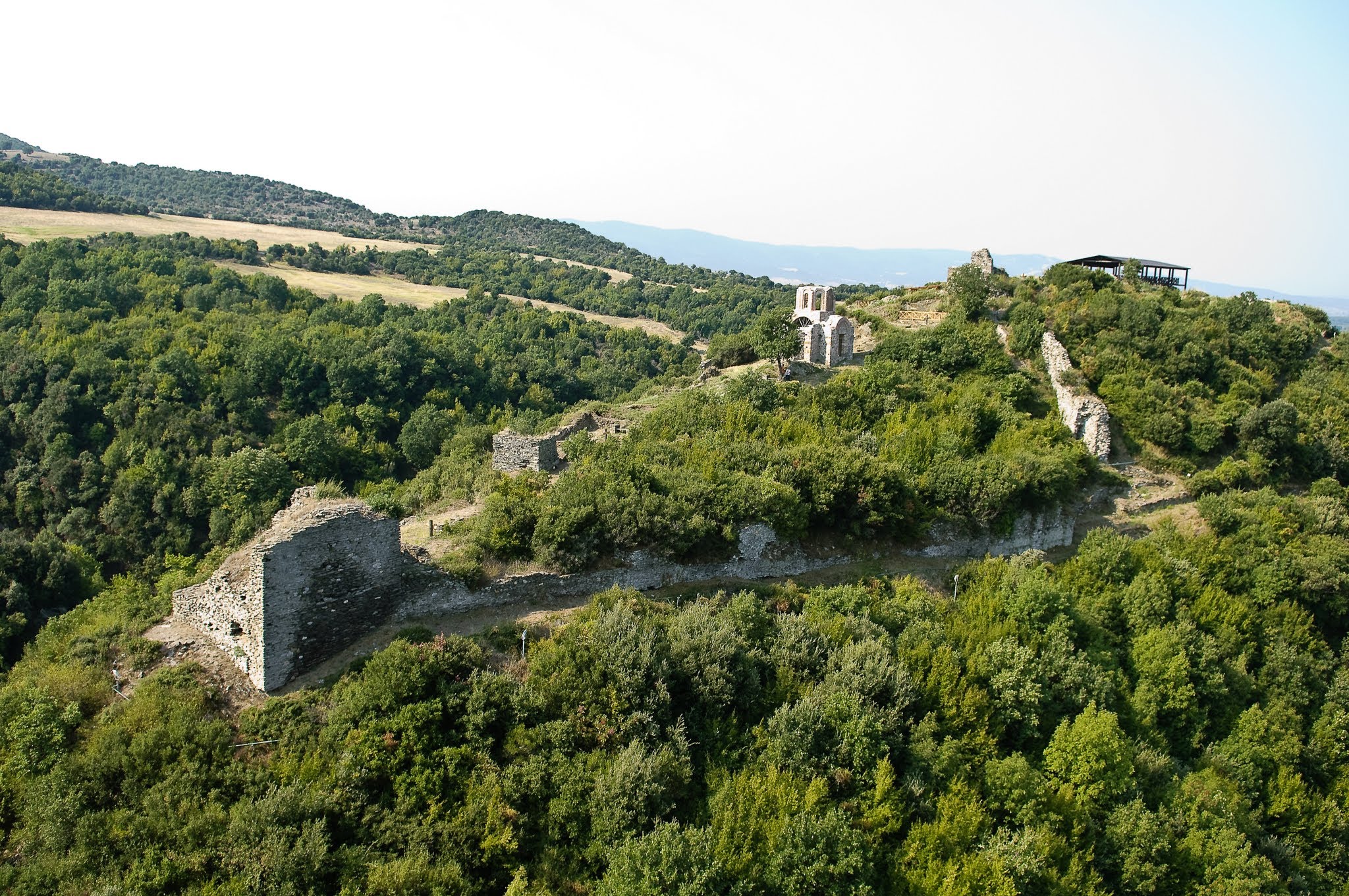 An aerial view of the stone fortifications and central church ruins of the Castle of Rentina, set against a backdrop of rolling green hills.