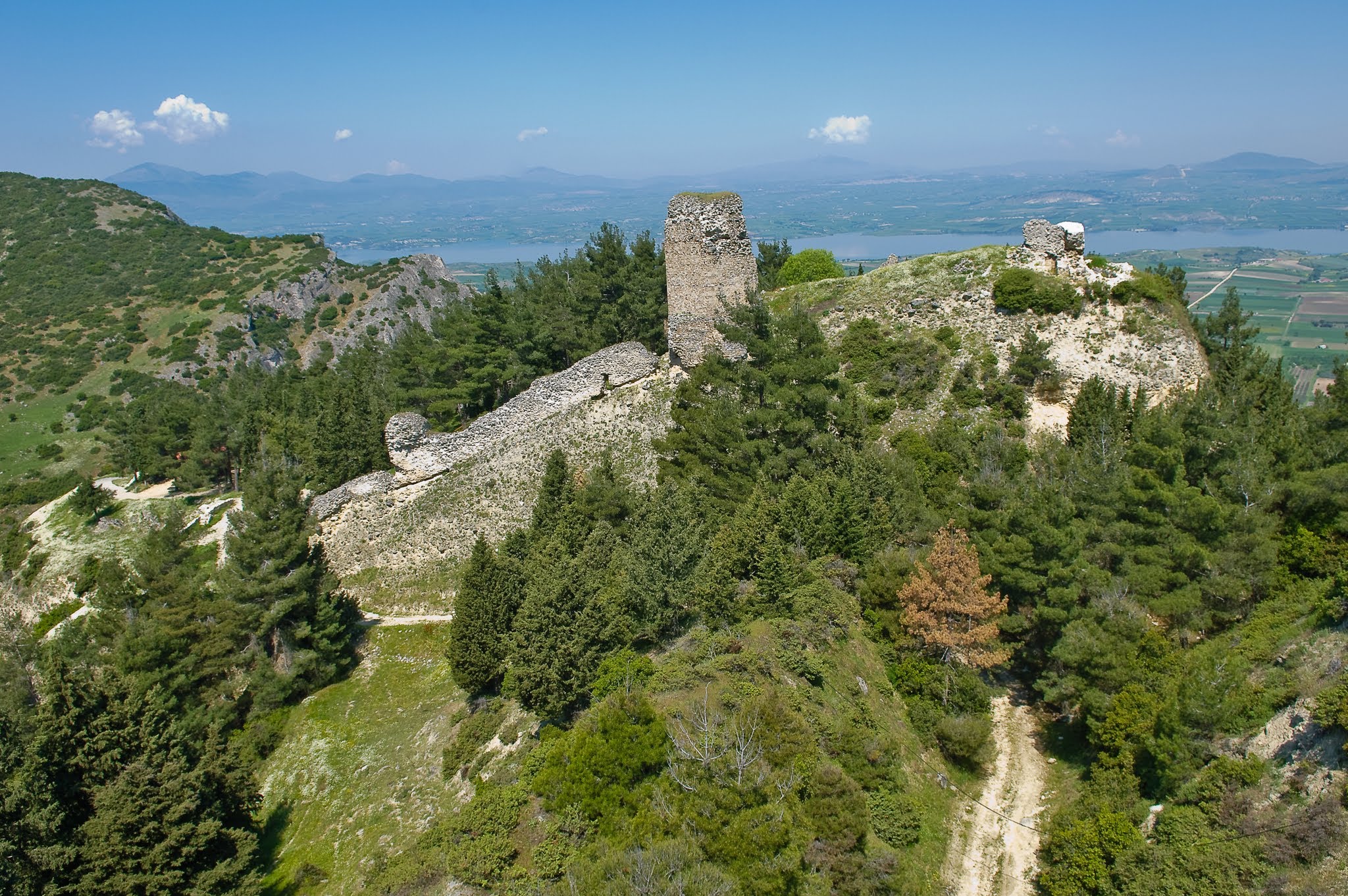 A wide oblique aerial photograph of the Byzantine Castle of Servia, showing the ruins of a tall central tower and defensive walls on a high ridge overlooking Lake Polyfytos.