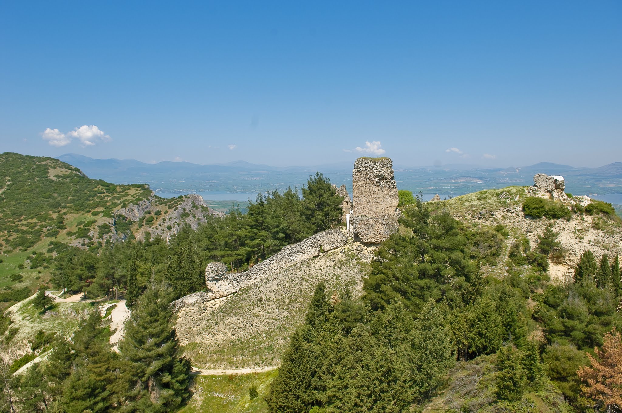 A wide oblique aerial view of the Castle of Servia ruins on a mountain ridge, featuring a tall Byzantine tower and fortification walls overlooking Lake Polyfytos.