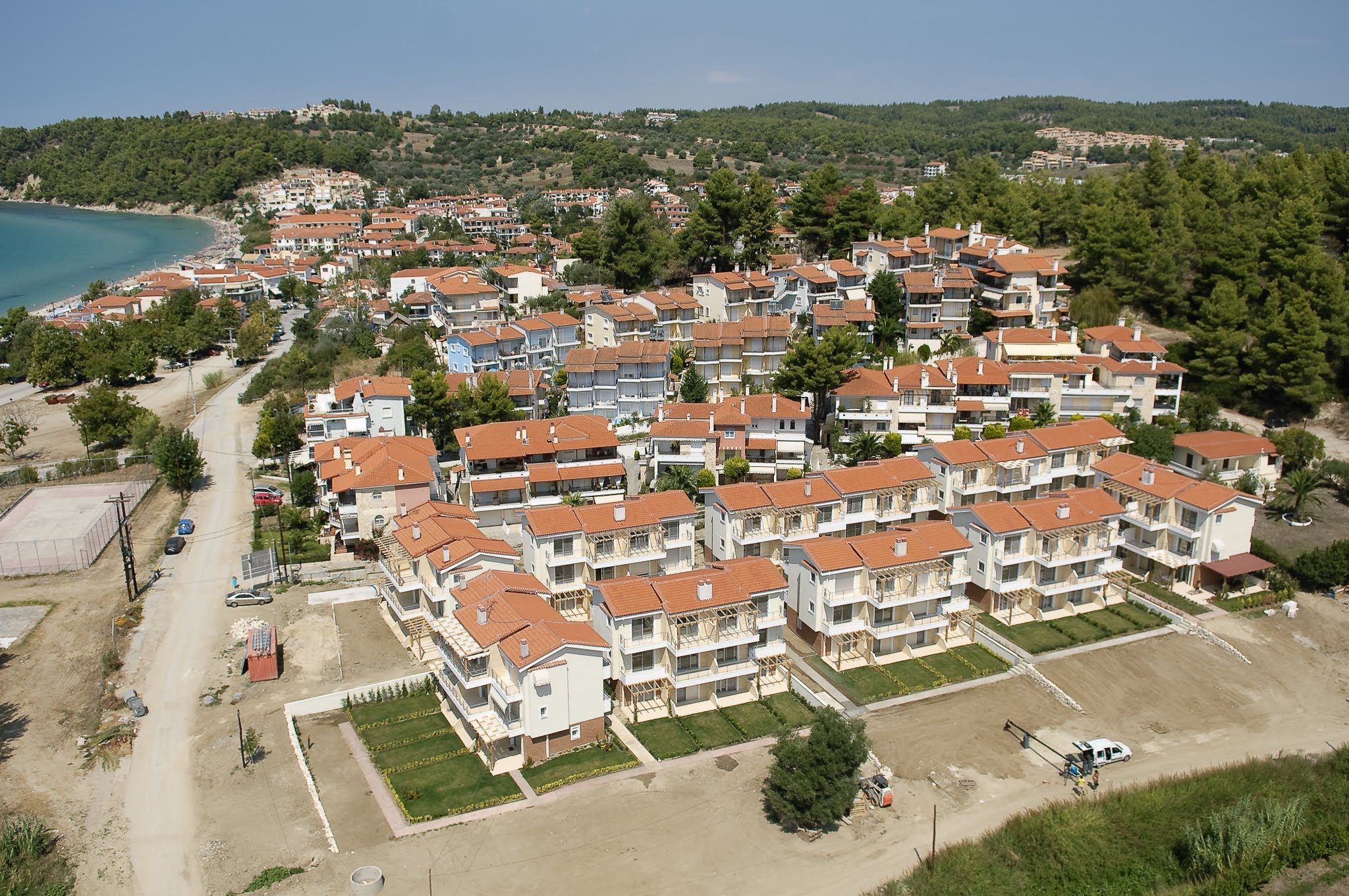 High-angle aerial view of a seaside residential community with terracotta-roofed villas, bordering a sandy bay and nestled against a pine forest.
